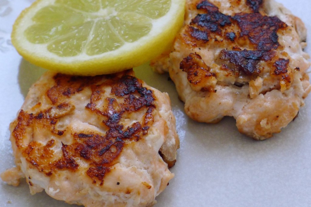 Two salmon fishcakes shown in close up with a lemon slice balancing on top in the top right of the frame. The fishcakes are shown whole and cooked with appealing crispy bits.