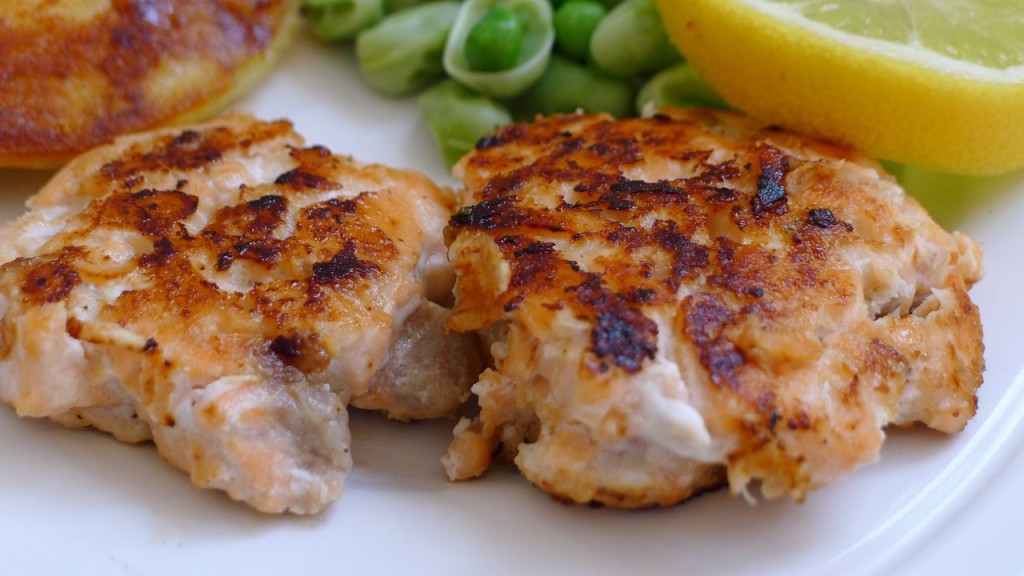 Two salmon fishcakes are shown close up in the foreground of the landscape frame. Other food on the plate is just visible in the background including peas and potatoes. A lemon slice is shown in the top rght. 