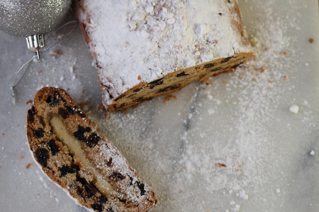 Overhead shot of a slice of gluten free stollen showing the fruits inside and marzipan layer., The remainder of the loaf is show with its dusting of sugar. 