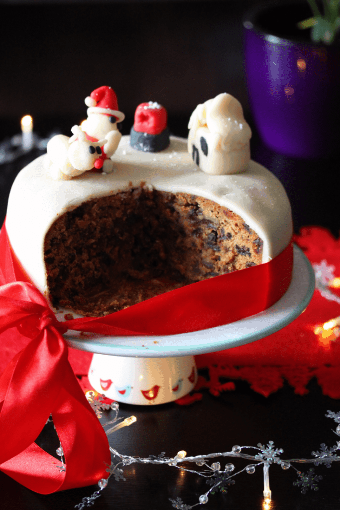 A close up of an iced Dairy Free Christmas Cake on a stand and surrounded by festive decorations. Several slices have been removed to show the dark fruity interior of the cake. 