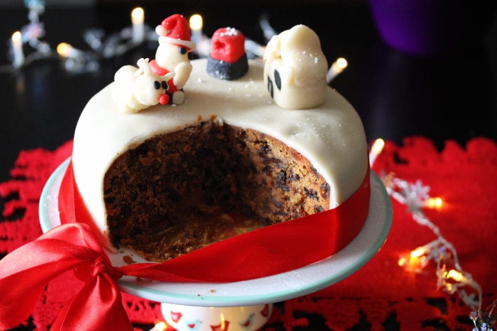 An iced Gluten Free Christmas Cake on a stand and surrounded by festive decorations. Several slices have been removed to show the dark fruity interior of the cake. 