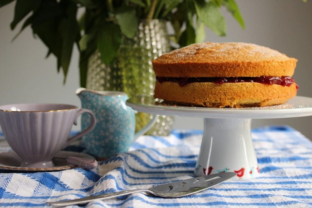 A table setting showing a victoria sponge on a cake stand to the right and a teacup and milk jug to the left. A cake cutter lays to the front. The shot is eye height at the middle jam layer. The cake is not cut. 