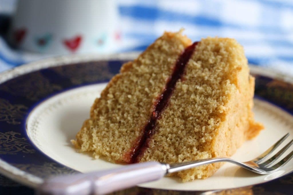 A slice of victoria sponge shown on a plate. The thin end of the wedge is towards the viewer with a fork laid down, handle again pointing to the viewer. You can see the underside of the sponge and middle jam layer. The background is out of focus. 