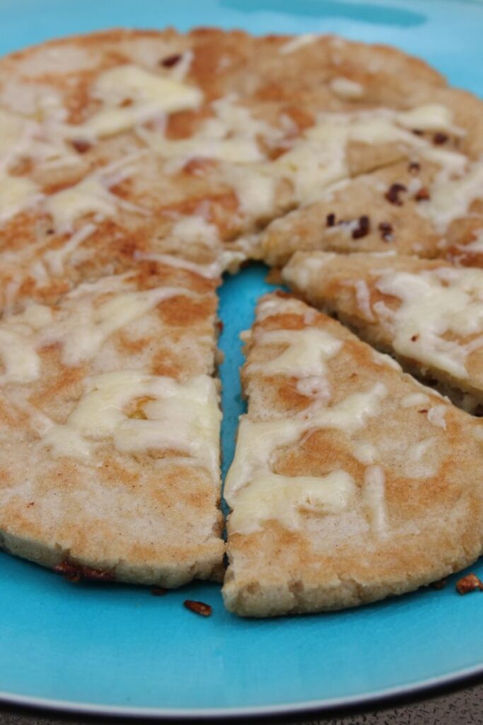 Gluten free garlic naan or flatbread on a blue plate with crispy piece of garlic scattered around. The garlic bread is topped with melted cheese.