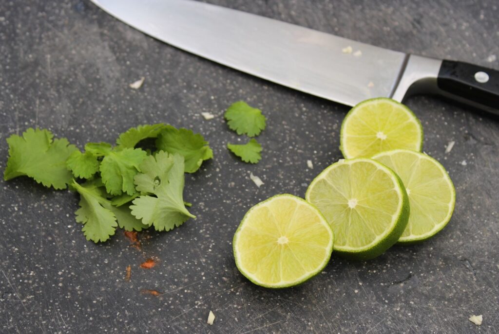 Slices of lime and coriander ready for chopping with a large sharp knife on a black chopping board.