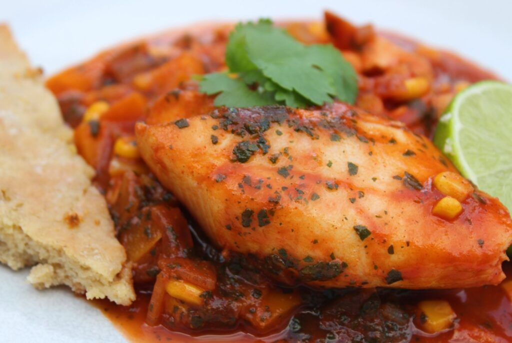 A fillet of fish in a spicy tomato sauce served with coriander and lime slice and a piece of garlic bread.